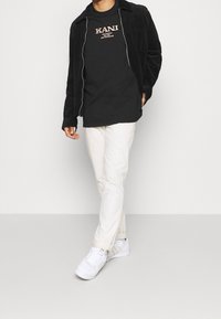 Černá kordová bunda přes černé tričko s potiskem, spárovaná s bílými kalhotami a bílými teniskami, na neutrálním pozadí.