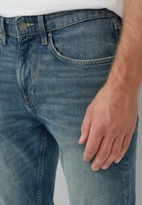 Jasnoniebieskie dżinsy o slim fit. Wyróżniają się kieszeniami z przodu, widocznymi szwami i zapięciem na guziki. Ręka opierająca się na udzie.