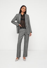 Grau kariertes Anzug mit tailliertem Blazer und geraden Hosen, kombiniert mit einer schwarzen, transparenten Bluse. Abgerundet wird das Outfit durch spitze beigen High Heels.