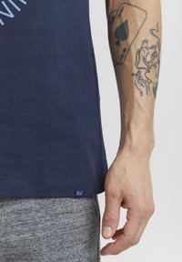 Tumed sinine T-särk pehmest puuvillast, millel on ümmargune kaelus ja väike logotag tähistust allservas. Hallid dressipüksid on nähtavad alt.