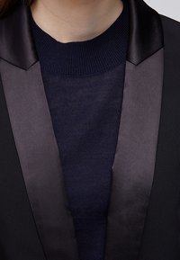 Svart kavaj med en slät satinrevers, bärs över en marinblå stickad tröja. Tyget är mjukt och framhäver kontrasten mellan texturer.