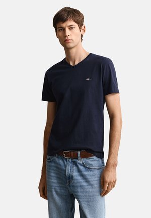 Mladić u tamnoplavoj V-izrez majici s malim logom i svijetloplavim trapericama s smeđim kožnim remenom, stoji ispred jednobojne pozadine.