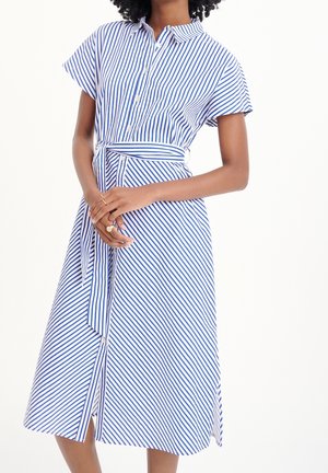 Vrouw met een blauw-wit verticale gestreepte korte mouw overhemdjurk met een bijpassende taille riem, handen zachtjes gevouwen voor zich.