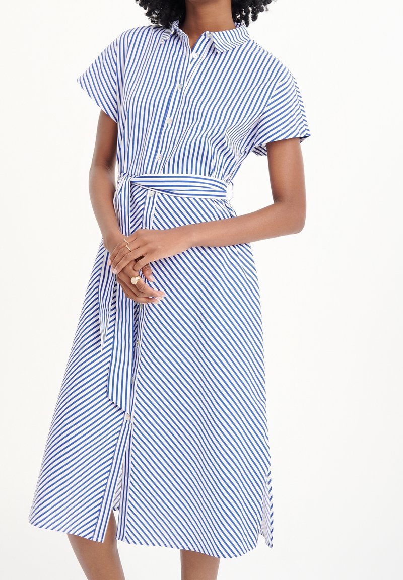 Femme portant une robe-chemise à manches courtes à rayures verticales bleu et blanc avec une ceinture assortie à la taille, mains doucement jointes devant.