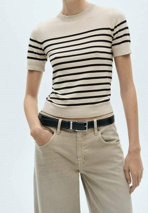 Beige korte mouw gebreide top met zwarte horizontale strepen gecombineerd met beige broek met hoge taille en een zwarte riem. Handen in de zakken.
