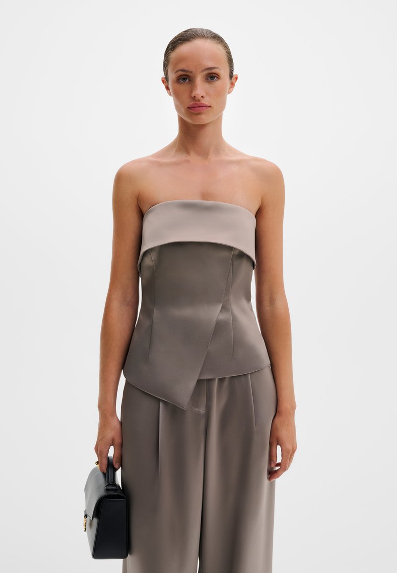 Γυναίκα που φοράει τοπ από σατέν σε τόνους ταφ με τιραντέ χωρίς λουράκια και παντελόνι με φαρδιά μπατζάκια, κρατώντας μια μικρή μαύρη τσάντα στο πλάι της.