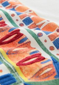 Close-up of colorful fabric with abstract geometric patterns in orange, blue, green, and red textured embroidery on white background.