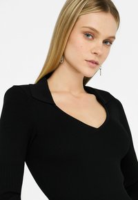Top nero a coste con scollatura a V profonda e colletto strutturato. Presenta maniche lunghe e un tessuto morbido e testurizzato che si adatta al corpo.
