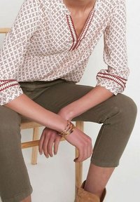 Blouse blanche à motifs géométriques rouges, col en V, manches retroussées. Pantalon court vert olive, bracelets dorés et bottes beige visibles.