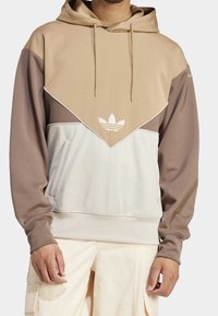 Muž v béžovo-hnědě-bílé blokované mikině s logem Adidas a stahovacími šňůrkami, ve spojení s krémovými cargo kalhotami.