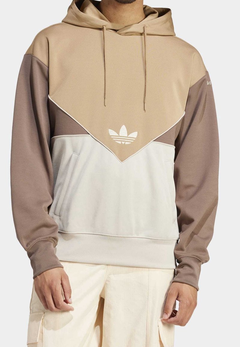 Muž v béžovo-hnědě-bílé blokované mikině s logem Adidas a stahovacími šňůrkami, ve spojení s krémovými cargo kalhotami.