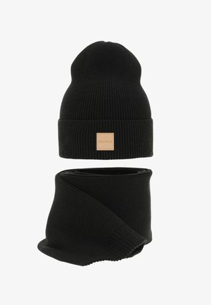 Bonnet en tricot noir avec une étiquette en cuir beige portant l'inscription « LESS IS MORE », assorti à une écharpe noire en tricot.