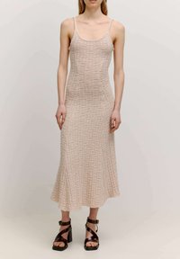 Strickkleid mit Spaghetti-Trägern, das ein strukturiertes, kariertes Muster in hellbeige zeigt. Fließendes, knielanges Design. Schwarze Riemchenheels.