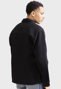 Black waffle-textured shirt with a collar, button cuffs, and side slit pockets. The design features a relaxed fit and a textured finish.