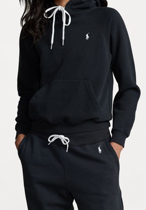 Person iført sort hættetrøje og matchende joggingbukser med hvide snore og lille hvidt hestelogo på brystet og låret.