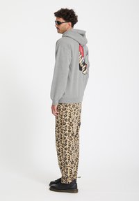 Homme portant des lunettes de soleil, un sweat à capuche gris avec un motif au dos, un pantalon imprimé léopard et des bottes noires, debout de profil sur fond blanc.