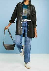 Svart läderjacka över en blå grafisk t-shirt, kombinerad med ljusblå vida jeans och vita sneakers. Grå axelväska ingår.