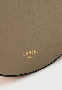 Στρογγυλή τσάντα από δέρμα ταουπέ με υφή. Διακοσμείται με χρυσό λογότυπο "LANCEL PARIS" και μαύρη λεπτομέρεια κατά μήκος της άκρης.