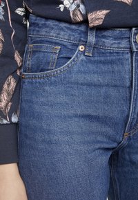 Vaqueros de mezclilla en azul medio con costuras naranjas. Presentan bolsillos frontales y un cierre de botón en la cintura. Top adyacente con estampado floral.