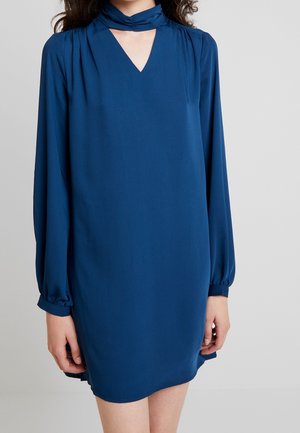 Vrouw die een blauwe jurk met lange mouwen tot op de knie draagt, met een V-hals en een bijpassende neksjaal tegen een egale achtergrond.