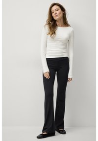 Langärmliges, weißes, eng anliegendes Oberteil mit gerafftem Detail an der Taille, kombiniert mit schwarzen, ausgestellten Hosen, beide aus glattem, elastischem Stoff. Schwarze flache Schuhe.