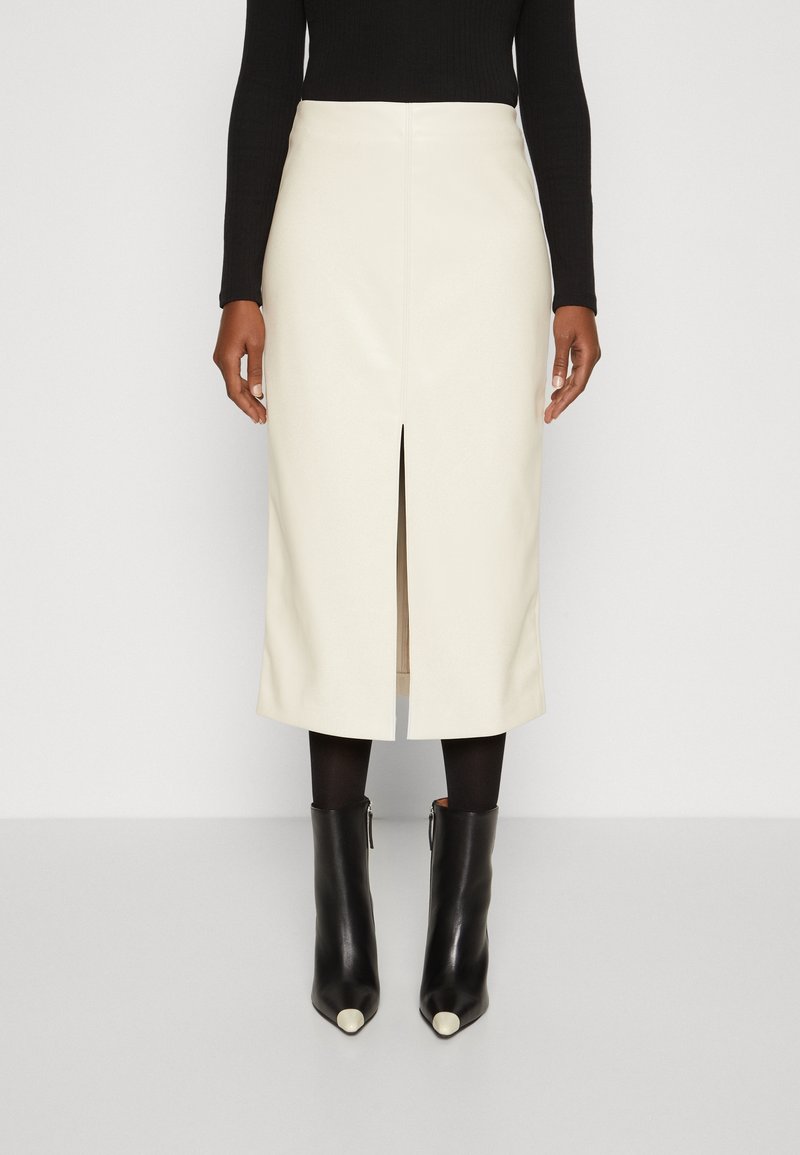 Femme portant une jupe midi blanche fendue devant, un haut à manches longues noir, des collants noirs et des bottines noires avec des bouts blancs.