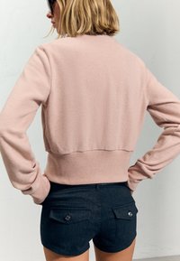 Roza skrajšana jopica z rebrastimi manšetami in robom, v kombinaciji s temnimi jeans kratkimi hlačami, ki imajo gumbaste zadnje žepe in prilegajočo se zasnovo.