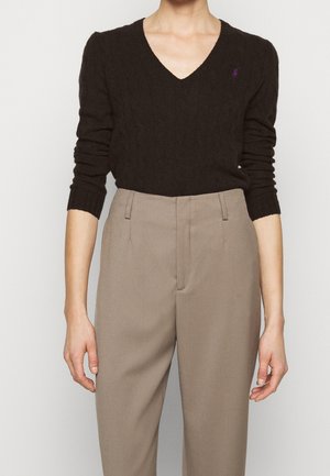 Personne portant un pull noir à col en V en maille torsadée et un pantalon beige taille haute, debout, les bras détendus le long du corps.