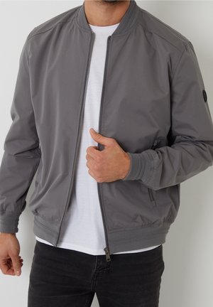 Mann trägt eine graue Bomberjacke über einem weißen T-Shirt, mit der Hand, die den Reißverschluss der Jacke nahe der Brust hält.