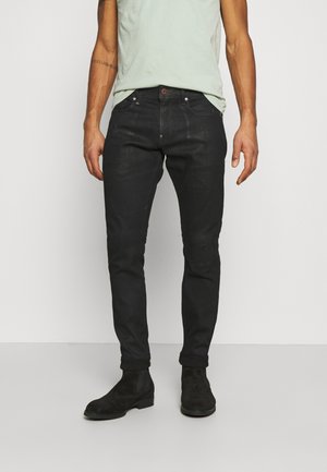 Mężczyzna w czarnych jeansach slim fit, czarnych skórzanych butach i jasnozielonym t-shircie, stojący na jednolitym białym tle.