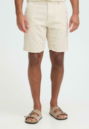 Shorts en lin beige à coupe droite, avec une fermeture à boutons et des poches latérales. Portés avec des sandales en daim marron.
