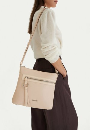 Donna che indossa un maglione color crema e pantaloni scuri con una borsa a tracolla beige dotata di una zip frontale e dettaglio con nappina.