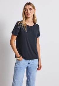 Top corto de manga corta gris oscuro con escote redondo y detalle de ojo de cerradura. Tejido suave, corte relajado, combinado con vaqueros de mezclilla azul claro.