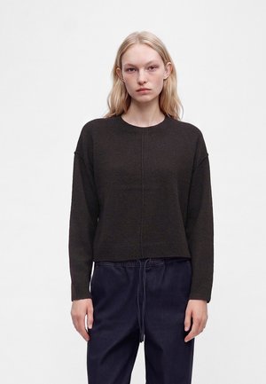 Jeune femme aux longs cheveux blonds portant un pull marron foncé à manches longues et un pantalon bleu foncé sur un fond clair uni.