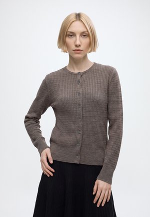 Jeune femme aux cheveux blonds courts porte un cardigan marron texturé boutonné et une jupe plissée noire, debout devant un fond blanc uni.