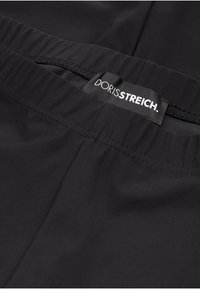 Ceinture en tissu noir avec l'étiquette logo "DORISSSTREICH." Elle présente une texture lisse et une bande élastique pour un ajustement adaptable.