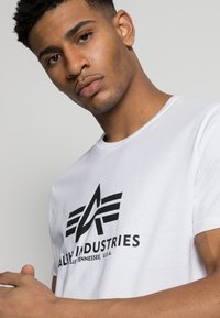 T-shirt alb din bumbac, cu un logo negru format din linii unghiulare și text, design cu guler rotund, cu o textură netedă și un stil minimalist.