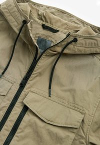 Veste légère couleur kaki avec une fermeture éclair à l'avant, une capuche ajustable et deux grandes poches. Tissu texturé avec une fermeture éclair et des cordons de tirage noirs contrastants.