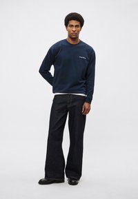 Tmavomodrá mikina s okrúhlym výstrihom a vyšívaným logom, v kombinácii so širokými tmavými rifľami a čiernymi topánkami. Hladká textúra a pohodlný strih.