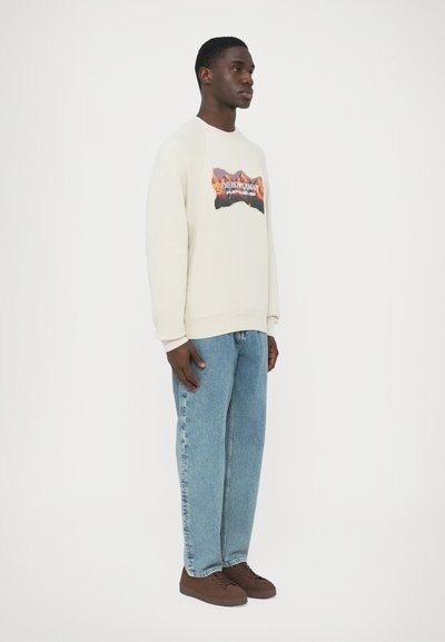 Kremowa bluza z nadrukiem, zestawiona z jasnoniebieskimi, luźnymi dżinsami i brązowymi trampkami. Bluza ma prążkowany kołnierz i mankiety.