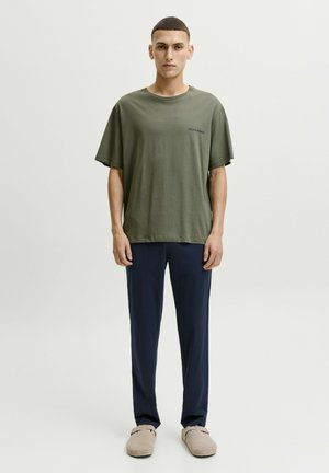 Young man standing wearing an olive green t-shirt, dark blue pants, and beige slip-on shoes against a plain light background.