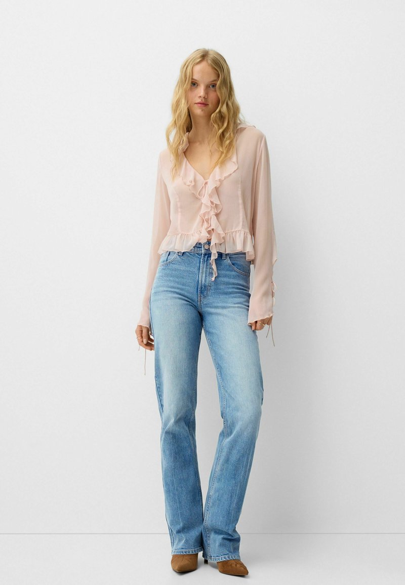Helltrosa Bluse mit Rüschensaum und langen Ärmeln, kombiniert mit hellblauen Schlagjeans und braunen Knöchelstiefeln. Glattes, leichtes Material.