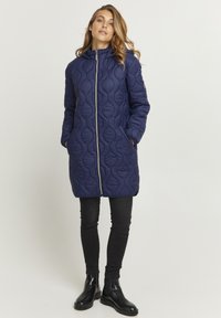 Cappotto lungo imbottito blu navy con zip frontale, cappuccio e due tasche laterali, caratterizzato da un motivo testurizzato e dettagli neri.