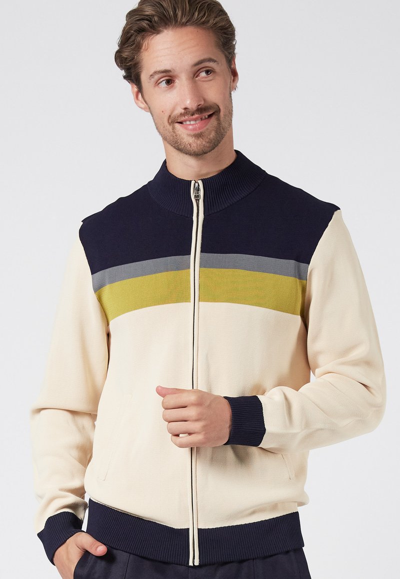 Zip-up jacka i krämvit med marinblå krage och ärmslut. Har horisontella ränder i grått och olivgrönt över bröstet. Texturerad tyg.