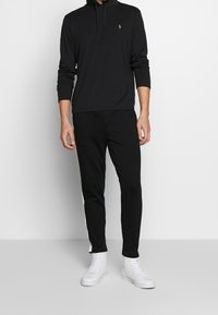 En person står klädd i en svart hoodie och matchande joggers, i kombination med vita högskaftade sneakers, mot en neutral bakgrund.