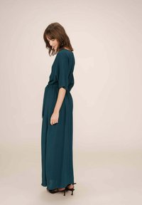 Lange teal jurk met een losse pasvorm, korte mouwen, een riem op de taille en een vloeiende silhouet. Draag met zwarte hoge hakken.