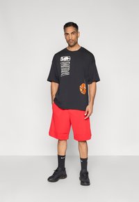 Μαύρο graphic t-shirt NBA με λευκές και πορτοκαλί λεπτομέρειες, σε συνδυασμό με κόκκινα σορτς μέχρι το γόνατο και μαύρα αθλητικά παπούτσια.