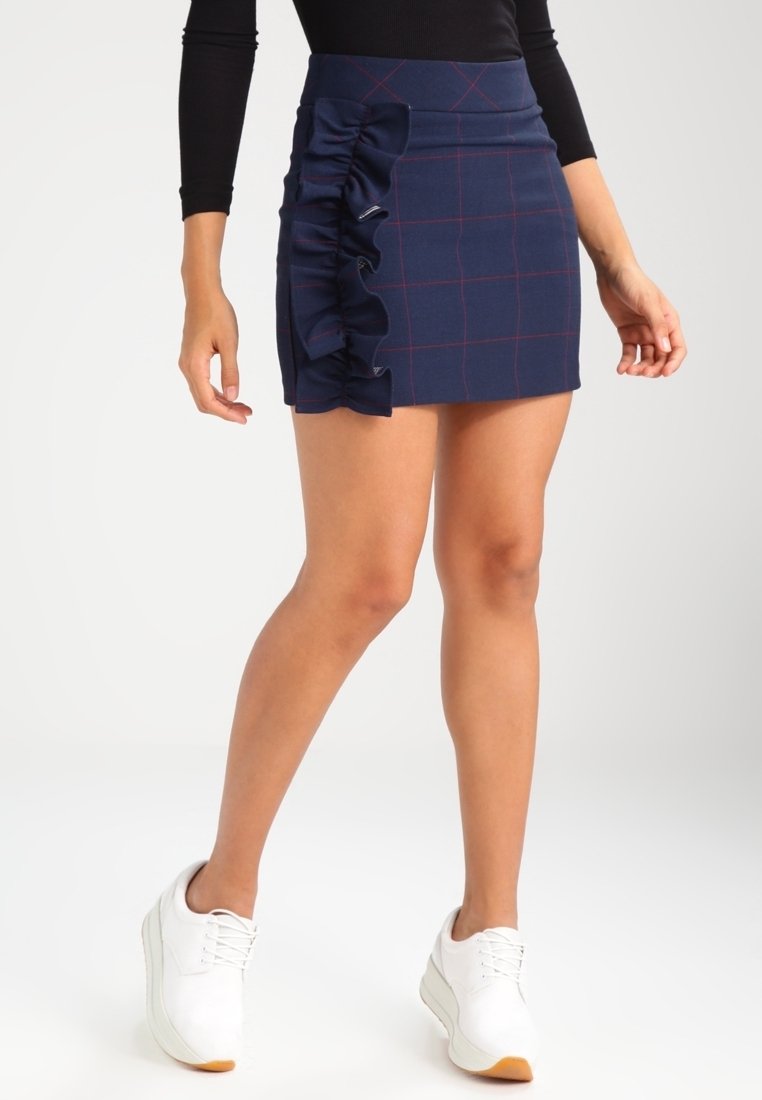 Mini-jupe bleu marine à motif quadrillé, avec un détail à volants sur le côté. Confectionnée dans un tissu lisse, associée à des baskets blanches pour un contraste.