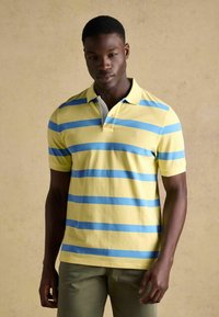 Jauns vīrietis stāv, valkājot dzeltenu polo kreklu ar zilām horizontālām svītrām un olīvu zaļas krāsas bikses uz vienkārša bēša fona.