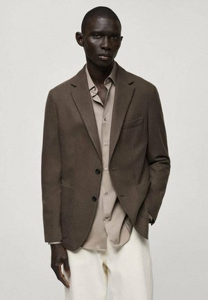 Blazer marrone su misura realizzato in tessuto liscio, con revers a notched, due tasche frontali e bottoni neri. Indossato sopra una camicia beige chiaro.